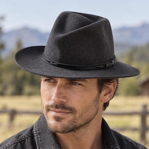 May include: A charcoal gray felt fedora hat featuring a pinched crown and a wide, structured brim. The hat is accented with a thin, black leather band that wraps around the base of the crown, secured with a small, rectangular metal detail.