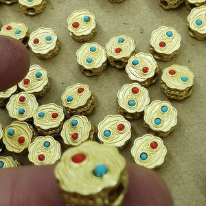 May include: Gold-coloured decorative beads with a floral design. Each bead features two red and two turquoise-coloured stones. The beads are small, with a detailed, textured surface. The beads are likely for jewellery making.