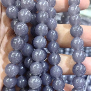 Pode incluir: Um fio de contas redondas de pedra preciosa de cor púrpura clara. As contas são lisas e têm um brilho ligeiro.