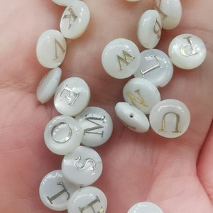 Puede incluir: Una colección de cuentas redondas blancas con letras doradas que deletrean el alfabeto. Las cuentas tienen aproximadamente 1 cm de diámetro.