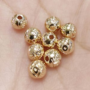 May include: A collection of gold-coloured, spherical beads with detailed embossed patterns. Each bead features a central hole for threading. The beads are shown against a soft background, emphasising their metallic shine and intricate design.