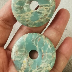 May include: Two turquoise-colored, donut-shaped stones with a hole in the center. The stones have a marbled appearance with red and cream-colored veins. The stones are held in a person's hand.