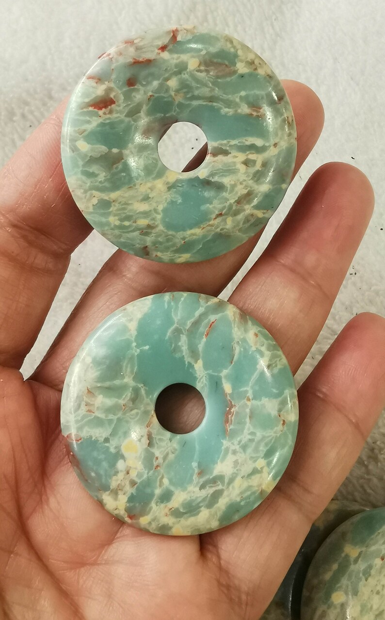 May include: Two turquoise-colored, donut-shaped stones with a hole in the center. The stones have a marbled appearance with red and cream-colored veins. The stones are held in a person's hand.