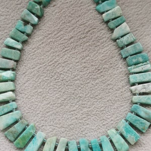 Puede incluir: Una hebra de cuentas de amazonita verde claro, cada cuenta tiene forma rectangular con un borde ligeramente irregular. Las cuentas están enhebradas en un hilo fino y transparente.