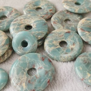 May include: A collection of donut-shaped, polished gemstones in varying sizes. The stones have a marbled appearance with shades of teal, cream, and red. The gemstones are likely used for jewelry making or other crafts.