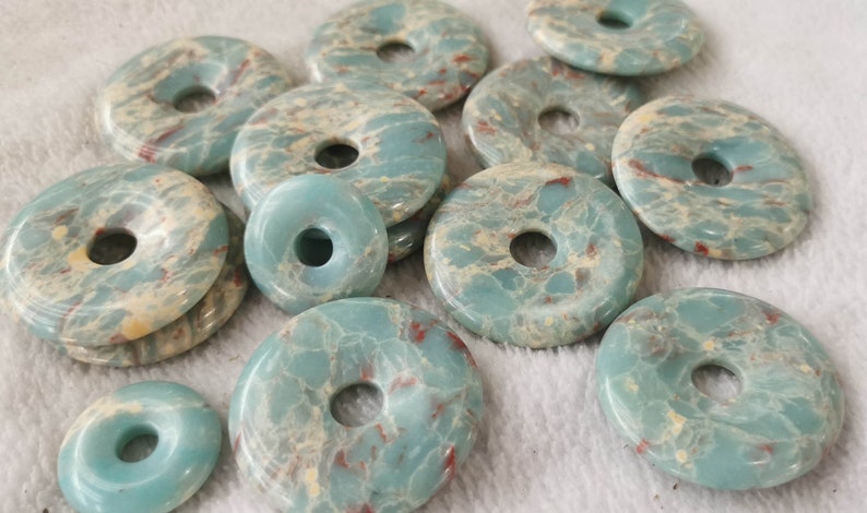 May include: A collection of donut-shaped, polished gemstones in varying sizes. The stones have a marbled appearance with shades of teal, cream, and red. The gemstones are likely used for jewelry making or other crafts.