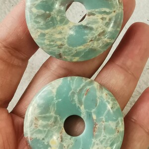 May include: Two round, donut-shaped stones with a hole in the center. The stones are a mix of light blue, cream, and reddish-brown colors. The stones have a marbled appearance and are likely used for jewelry or crafts.
