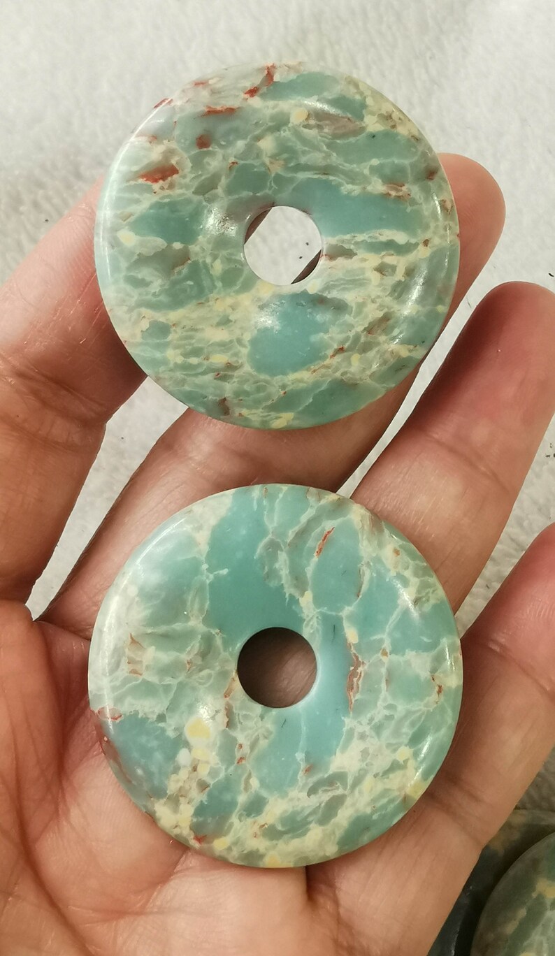 May include: Two round, donut-shaped stones with a hole in the center. The stones are a mix of light blue, cream, and reddish-brown colors. The stones have a marbled appearance and are likely used for jewelry or crafts.