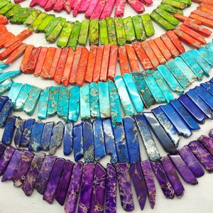 May include: A collection of colorful, patterned, rectangular gemstone beads. The beads are arranged in a fan shape, with the colors transitioning from pink to green to orange to blue to purple.