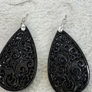 Könnte beinhalten: Ein Paar schwarze Hängeohrringe mit einem silbernen Haken. Die Ohrringe sind aus einem geschnitzten Material mit einem wirbelnden Muster.