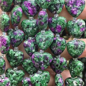 May include: Close-up of a strand of heart-shaped beads. The beads are a mix of green and purple, with a speckled pattern. Each bead is approximately 2.5 cm in diameter. The beads are strung together with a clear string, creating a necklace or bracelet.