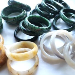 Peut inclure: Une collection de bracelets en jade et agate dans différentes nuances de vert, blanc et jaune. Les bracelets sont empilés et disposés sur une surface blanche.