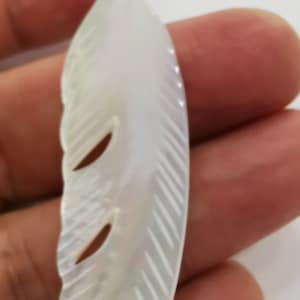 May include: A white mother of pearl feather pendant with a detailed carved design. The pendant is shaped like a feather with a pointed tip and a rounded base.