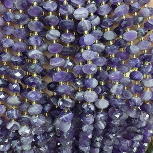 Pode incluir: Um fio de contas de ametista facetadas, cada conta é de cor roxo escuro com veios brancos. As contas são enfiadas em um fio de ouro.