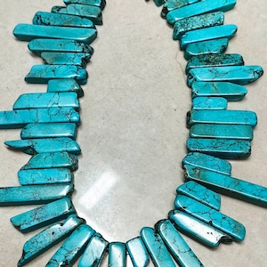 Puede incluir: Una hebra de cuentas de piedra turquesa, cada cuenta es de forma rectangular larga y delgada, con un acabado rugoso y natural. Las cuentas son de un color azul verdoso vibrante con vetas y marcas marrones.