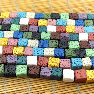 May include: A collection of colourful lava rock beads, each bead is a small cube with a rough, textured surface. The beads are arranged in a row, showcasing a variety of colours, including blue, green, red, yellow, and white.