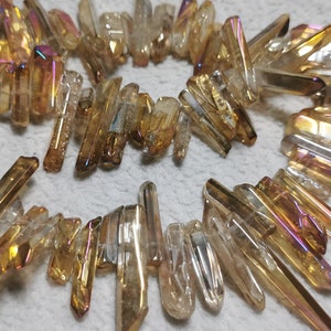 Może przedstawiać: Szereg iryzujących złotych i przezroczystych punktów kryształu kwarcowego. Punkty mają około 2,5 cm długości i mają fasetowany, spiczasty kształt.