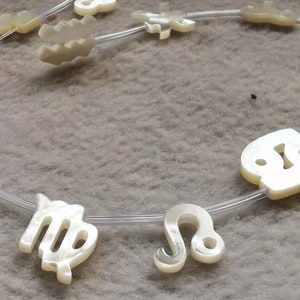 Könnte beinhalten: Eine Halskette mit Tierkreiszeichen in einer hellen, perlmuttartigen Farbe. Die Symbole sind auf einem transparenten, flexiblen Band aufgereiht. Die Halskette wird auf einer strukturierten, hellgrauen Oberfläche präsentiert.