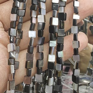 May include: Strands of square beads in shades of black, gray, and white. The beads are strung together, creating a necklace or bracelet. The beads have a pearlescent sheen, adding a touch of elegance. The beads are approximately 0.25 inches in size.