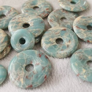 May include: A collection of turquoise-colored gemstone donut pendants with a hole in the center. The pendants have a marbled appearance with cream and red veining. The pendants are of varying sizes and are arranged on a white surface.
