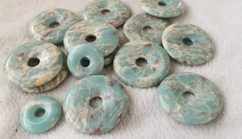 May include: A collection of turquoise-colored gemstone donut pendants with a hole in the center. The pendants have a marbled appearance with cream and red veining. The pendants are of varying sizes and are arranged on a white surface.