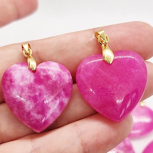 May include: Two heart-shaped pendants in shades of pink and magenta. The pendants have a polished, smooth surface with a marbled appearance. Each pendant has a gold-colored bail for attaching to a chain. The pendants are held in a hand.