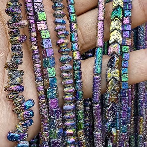 Pode incluir: Uma variedade de contas de pedras preciosas iridescentes em diferentes formas e tamanhos, incluindo quadradas, redondas e ovais. As contas têm um acabamento metálico com um arco-íris de cores, incluindo roxo, azul, verde e dourado.