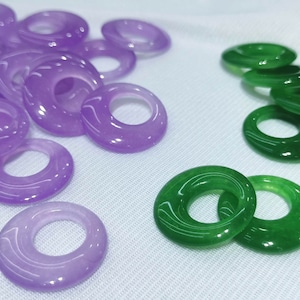 May include: A collection of purple and green jade rings. The rings are smooth and polished, with a hole in the center. They are arranged in a pile on a white surface.
