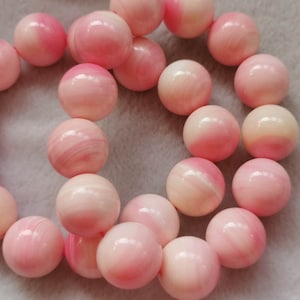 Könnte beinhalten: Eine Kette aus rosa und weißen Muschelkugeln, wahrscheinlich aus Conchenschalen, die in einem Kreis angeordnet sind. Die Perlen sind glatt und poliert und haben ein natürliches, organisches Aussehen.