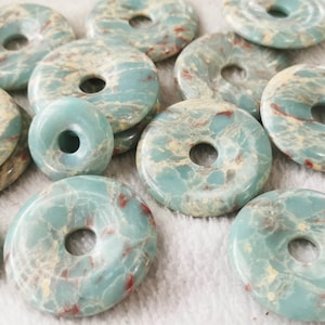 May include: A collection of turquoise-colored gemstone donut beads with a central hole. The beads have a marbled appearance with cream and red veining. The beads are of varying sizes and shapes, suitable for jewelry making.