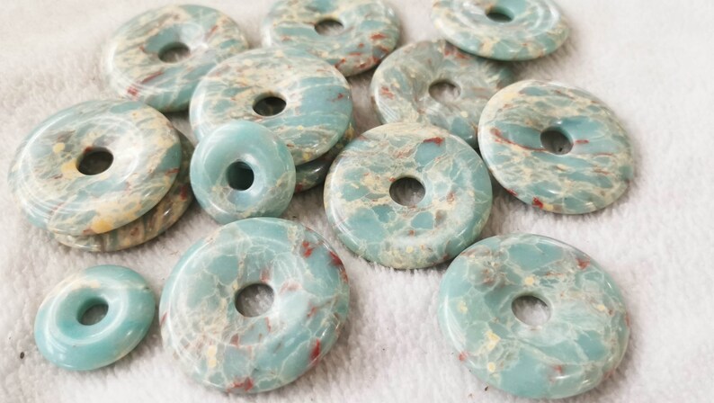 May include: A collection of turquoise-colored gemstone donut beads with a central hole. The beads have a marbled appearance with cream and red veining. The beads are of varying sizes and shapes, suitable for jewelry making.