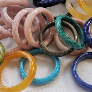 May include: A collection of colorful stone bangles in various shades of pink, green, blue, and yellow. The bangles are made of polished stone and have a smooth, glossy finish.