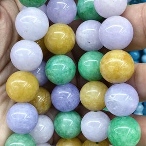 Pode incluir: Fios de contas redondas de pedras preciosas em várias cores, incluindo verde, amarelo e lavanda. As contas têm aproximadamente 1 cm de diâmetro, são enfiadas e seguradas em uma mão.