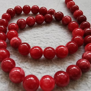 Può includere: Una collana di perle di pietre preziose rosse. Le perle sono rotonde e hanno una finitura liscia e lucida. Le perle sono infilate su un filo trasparente.