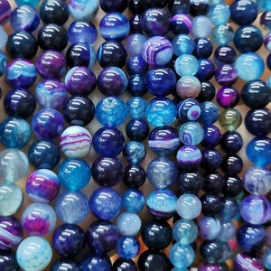 Pode incluir: Uma coleção de contas de ágata azul, roxa e branca. As contas são redondas e têm um acabamento liso e polido. São perfeitas para fazer joias ou outros trabalhos manuais.