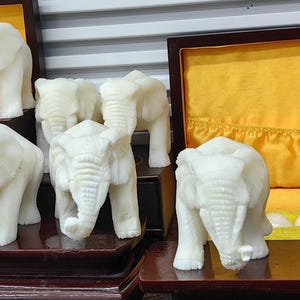 以下が含まれることがあります： 白い象の彫刻の置物コレクション。立っているものもあれば、座っているものもあります。置物は木製の棚と木製の箱に飾られています。