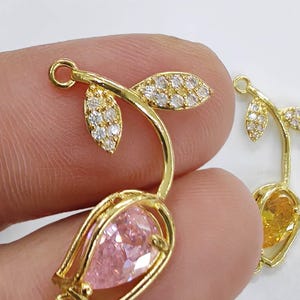 May include: Gold-tone flower charm with a pink pear-shaped stone and two leaves with clear stones. The charm has two loops for attaching to a necklace or bracelet.