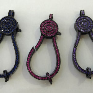 Peut inclure: Trois fermoirs en métal noir en forme de larme. Chaque fermoir est orné d'un cercle de strass colorés. Les fermoirs sont bleus, roses et violets.