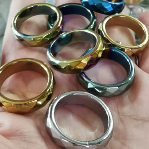 May include: A collection of seven iridescent gemstone rings. The rings are faceted and have a variety of colors, including gold, blue, and silver.