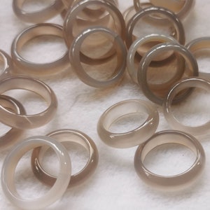 Puede incluir: Una colección de anillos de ágata gris, cada uno con un acabado liso y pulido. Los anillos están apilados y dispersos sobre una superficie blanca.