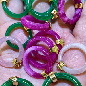 Könnte beinhalten: Eine Sammlung von Jade-Ringen mit goldenen Akzenten. Die Ringe sind in verschiedenen Farben, darunter Grün, Weiß und Lila. Die Ringe werden auf einer Hand präsentiert.