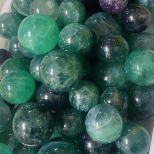 May include: A collection of green fluorite spheres of various sizes. The spheres are smooth and polished, with a translucent green color. Some of the spheres have a slight purple hue.