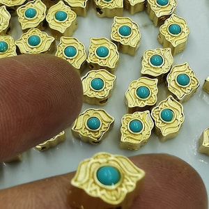 May include: A close-up of gold-coloured metal beads with turquoise gemstones. The beads have a decorative design and are scattered on a white surface.