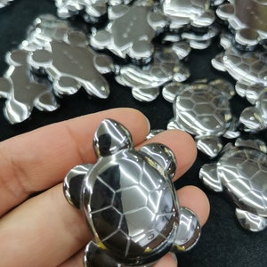 May include: A collection of shiny, silver-coloured turtle-shaped hematite stones. The stones are smooth and polished, with a reflective surface. The stones are arranged in a pile, with one stone in the foreground.