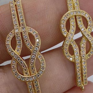 Pode incluir: Dois pingentes dourados em forma de símbolo do infinito, incrustados com pequenas pedras preciosas transparentes. Os pingentes têm um acabamento polido e são projetados com uma argola na parte superior para pendurar. As pedras preciosas são colocadas próximas umas das outras, criando um efeito brilhante.