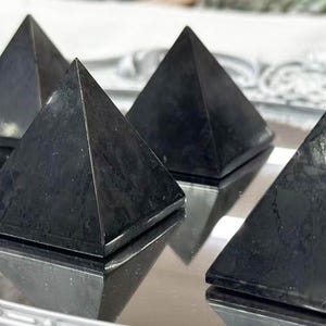 May include: Six black pyramid-shaped objects with a textured surface are arranged on a reflective silver tray. The pyramids have a geometric design and are evenly spaced, creating a symmetrical pattern.