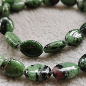 Puede incluir: Una hebra de cuentas de piedras preciosas ovaladas de color verde, negro y rosa. Las cuentas están enhebradas en un hilo transparente.