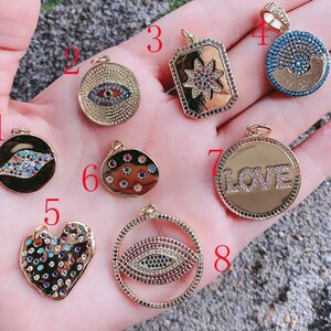 May include: A selection of gold-coloured pendants featuring diverse designs. The pendants showcase an eye, a starburst, a heart, and the word "LOVE". Each pendant is embellished with colourful gemstones and detailed craftsmanship, ideal for jewellery making.