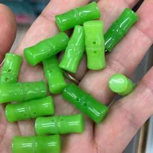 May include: A collection of bright green, bamboo-shaped beads. Each bead has a glossy finish and is cylindrical with carved details to mimic bamboo stalks. The beads are held in a hand, showcasing their vibrant color and design.