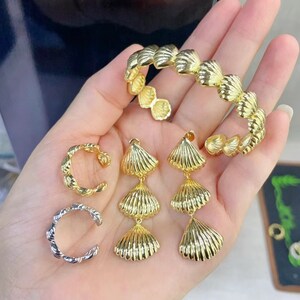 Pode incluir: Um conjunto de joias douradas com design de concha. O conjunto inclui uma pulseira, duas argolas para orelha e um par de brincos pendurados. Os brincos têm três níveis de pingentes em forma de concha.